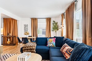 A modern living room with a sectional sofa, a dining area, and a large window with curtains. - South Bay Hideaway - 3BR/2BA Near LAX, Beaches & SoFi - Monthly Stays (Lawndale)