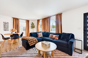 A modern living room with a dark blue sofa, a round coffee table, and a dining area with a white table and black chairs. - South Bay Hideaway - 3BR/2BA Near LAX, Beaches & SoFi - Monthly Stays (Lawndale)