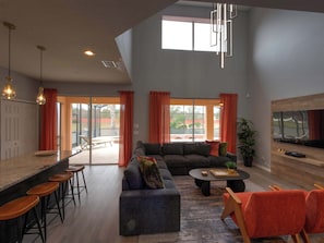 A spacious living area with a large sofa, a coffee table, and a bar with stools. There is a television mounted on the wall and a window providing natural light. - Palms & Pixie Dust Villa - Pool (Davenport)