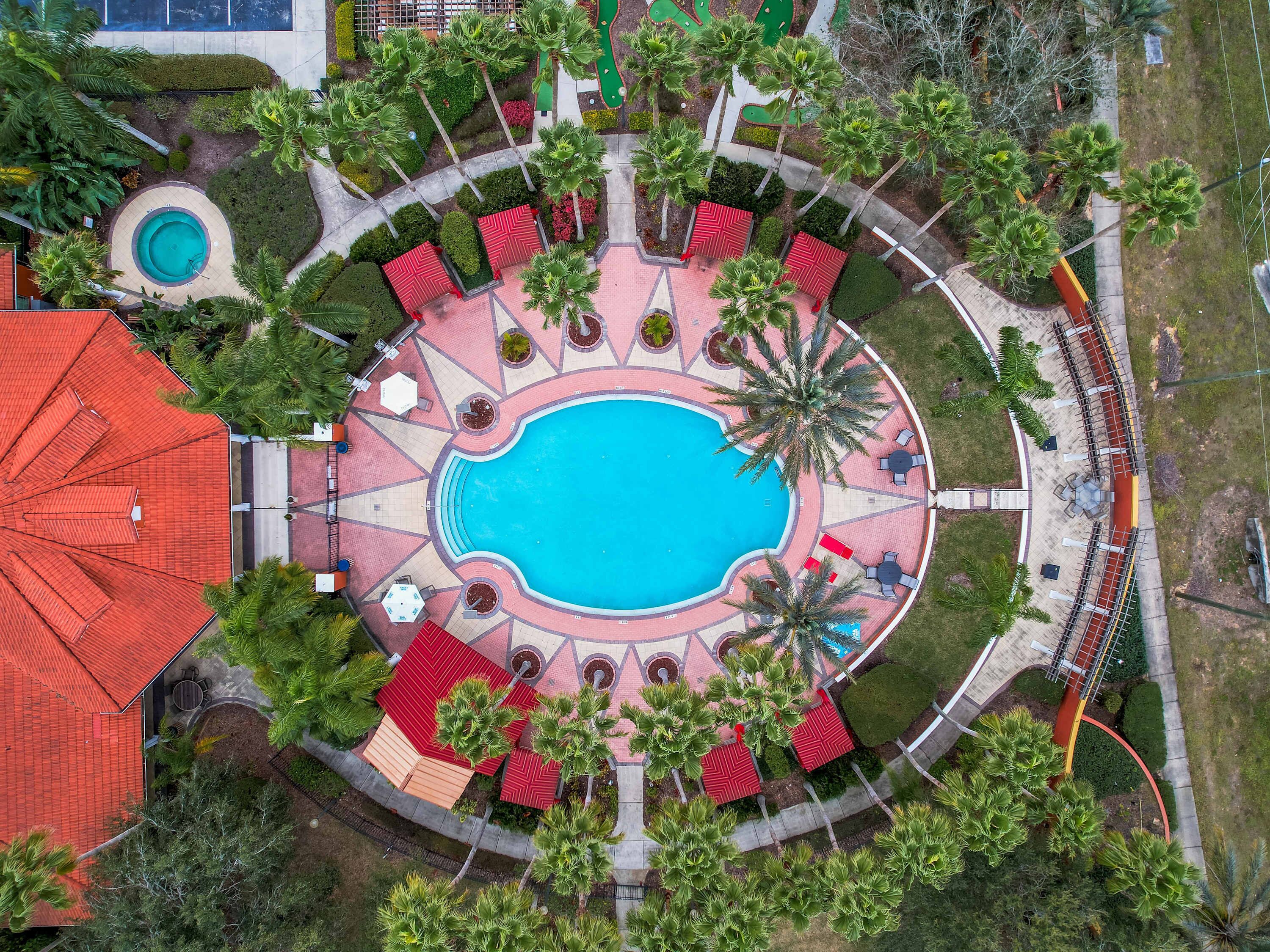 An aerial view of a resort with a central swimming pool, surrounded by lounge areas with red and white patterned flooring, and palm trees.