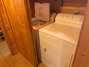 A compact laundry room with a washer and dryer, wooden door, and tiled floor. - Mountain Lake Retreat | Kayaks, Dock + Fire Pit (Mentone)
