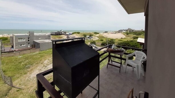 Uma varanda com churrasqueira, mesa e cadeiras, e vista para a praia e edifícios.