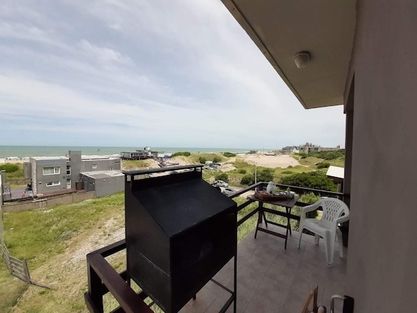 Un balcón con una parrilla, una mesa con sillas y vistas a la playa y edificios.