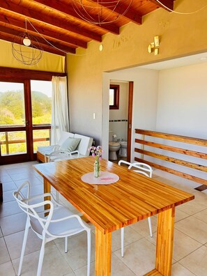 Una mesa de comedor de madera con sillas, un florero con flores y vistas a un balcón con baranda de madera.