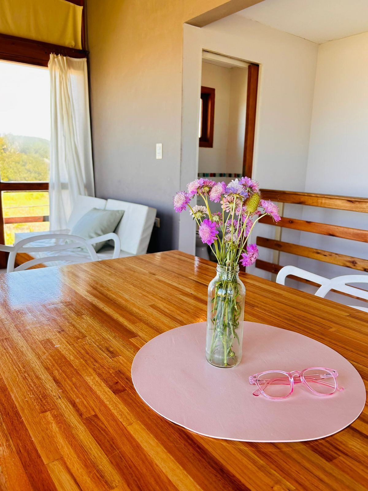 Una mesa de comedor de madera con un mantel individual rosa, un florero de vidrio transparente con flores y un par de gafas rosas sobre el mantel.