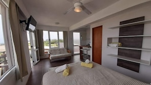 Quarto de hotel com cama, TV fixada na parede, ventilador de teto e vista para o exterior.