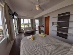 Una habitación de hotel con una cama, un televisor montado en la pared, un ventilador de techo y vistas al exterior.