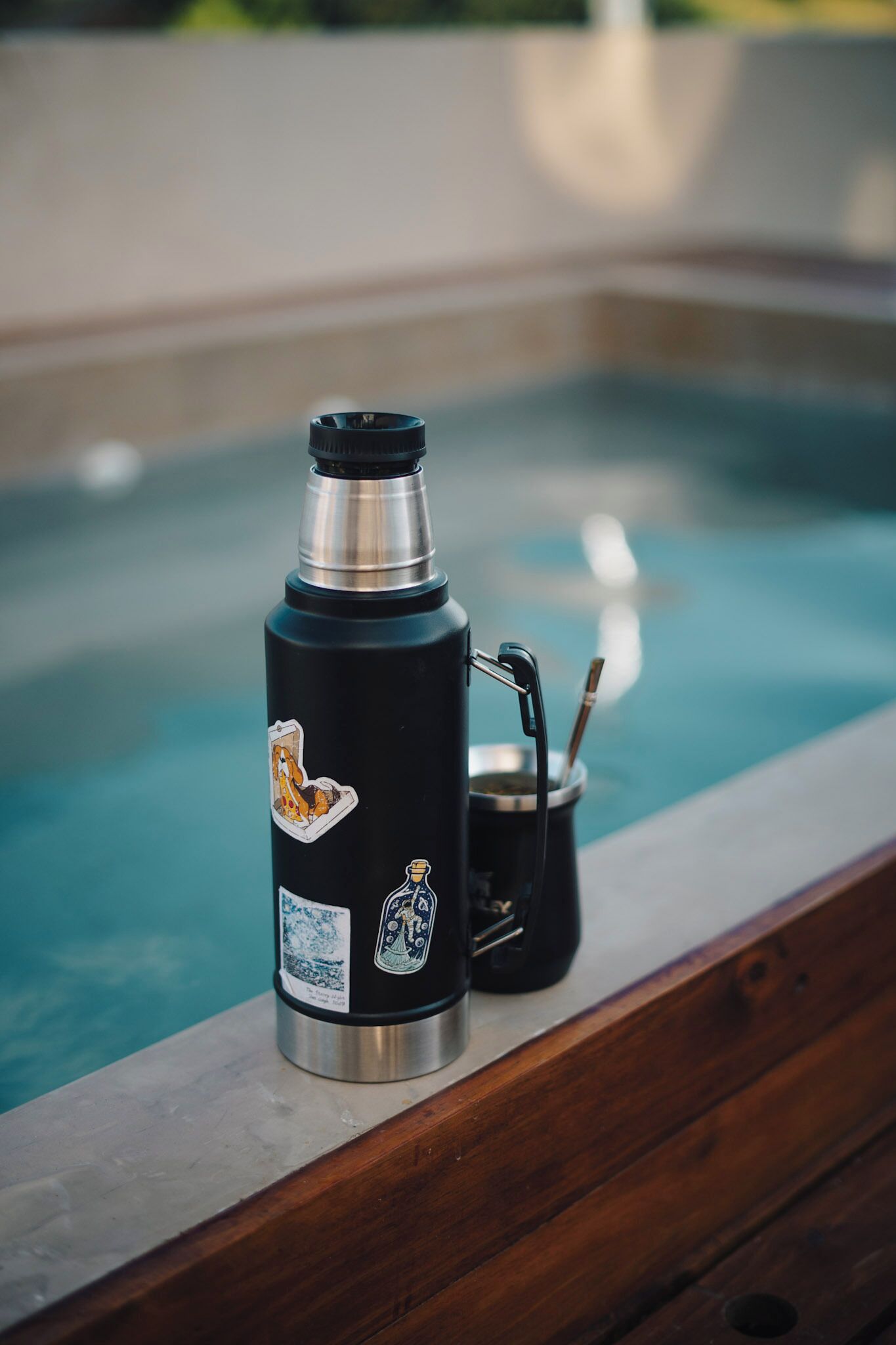A stainless steel thermos with stickers on a wooden ledge by a pool.