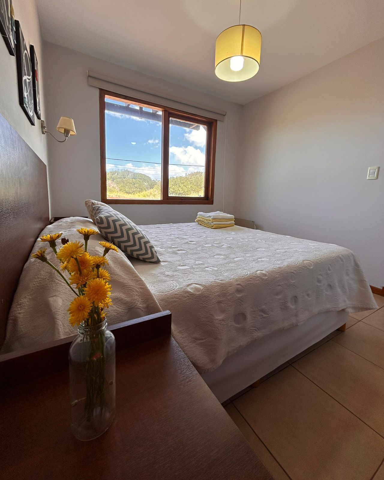 Un dormitorio con cama, una ventana con vistas, un florero con flores y una lámpara.