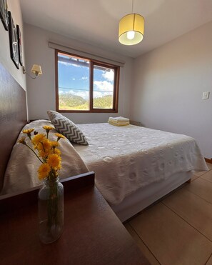 Une chambre à coucher avec un lit, une fenêtre donnant sur un beau paysage, un vase rempli de fleurs et une lampe.