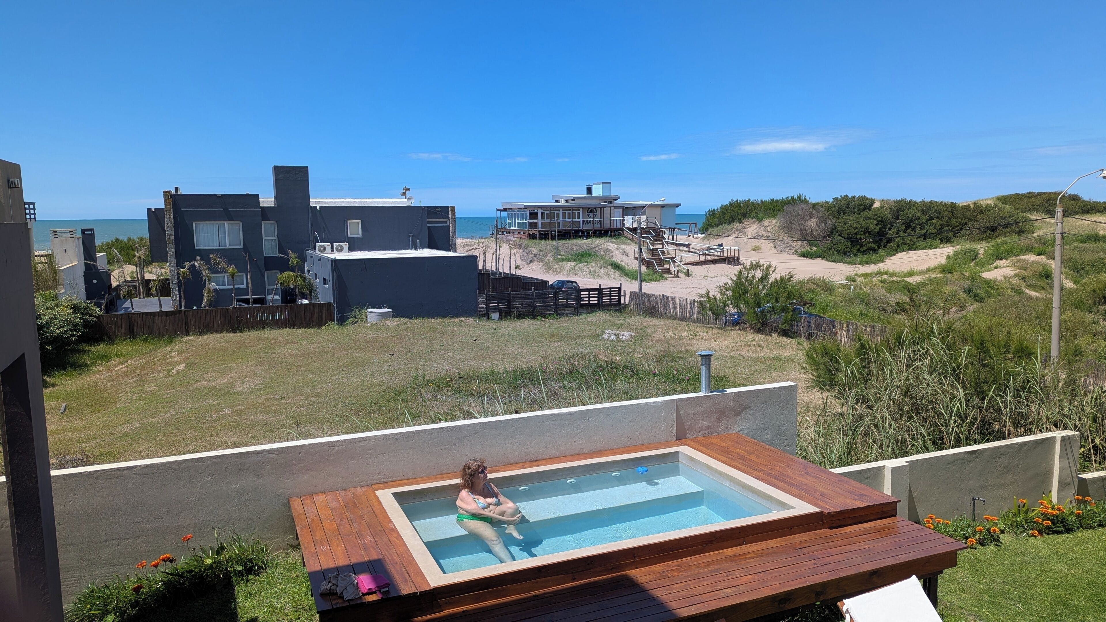 A swimming pool with a person, surrounded by a deck and a view of the beach and buildings.