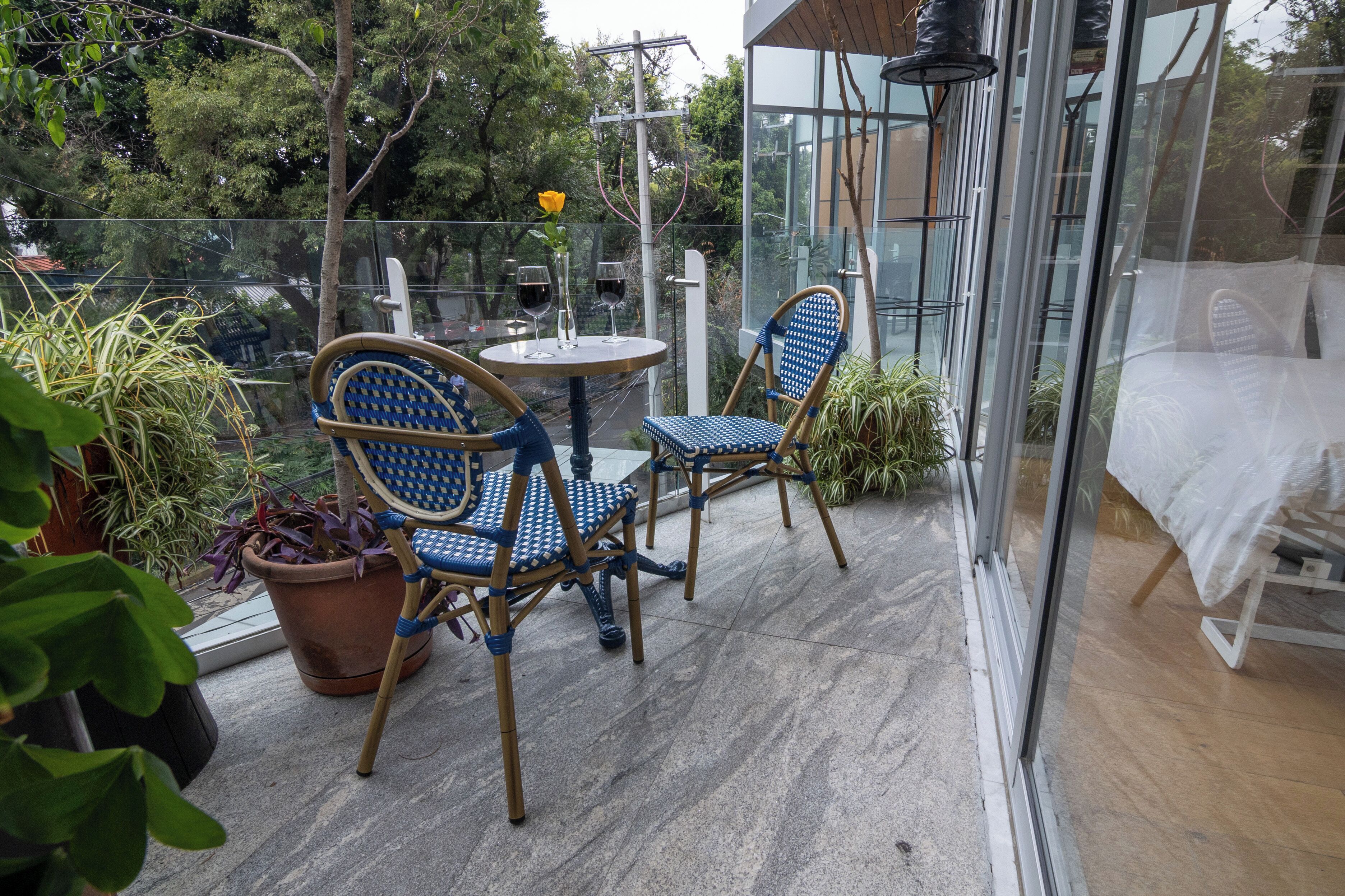 Ein gläserner Balkon mit zwei Korbstühlen und einem kleinen runden Tisch, umgeben von Blumentöpfen und einer gelben Blume.