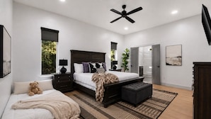 Quarto moderno com cama grande, mesa de cabeceira, abajur, ventoinha no teto, janela, porta, tapete e um urso de pelúcia.