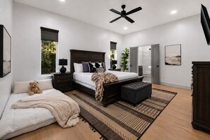A modern bedroom with a large bed, a bedside table, a nightstand, a floor lamp, a ceiling fan, a window, a door, a rug, and a teddy bear. - Luxury 7BR Cabin | Pickleball, Sauna & Hot Tub (Broken Bow)