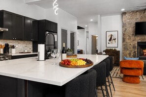 A modern kitchen with a large island, dark cabinets, and a stone fireplace. - Luxury 7BR Cabin | Pickleball, Sauna & Hot Tub (Broken Bow)