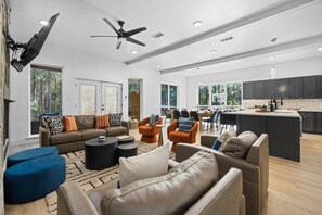 Een ruime woonkamer met een bank, fauteuils en een salontafel. Er is een eethoek met stoelen en een tafel, en een keuken met donkere kastjes en een aanrecht.