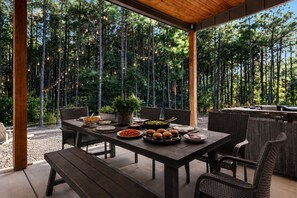 An outdoor dining area with a wooden table and chairs, surrounded by a bamboo fence, and a grill station to the right. - Luxury 7BR Cabin | Pickleball, Sauna & Hot Tub (Broken Bow)