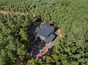 A house surrounded by dense forest, with a large metal roof and a tennis court. - Luxury 7BR Cabin | Pickleball, Sauna & Hot Tub (Broken Bow)