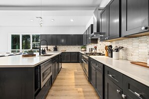 A modern kitchen with dark cabinetry, a large island, and a white backsplash. - Group Retreat | Hot Tub + Sauna | Sleeps 24 (Broken Bow)