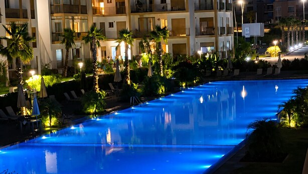 Ein Swimmingpool mit blau beleuchtetem Rand, umgeben von Palmen und Liegestühlen, mit einem Hotelgebäude im Hintergrund.