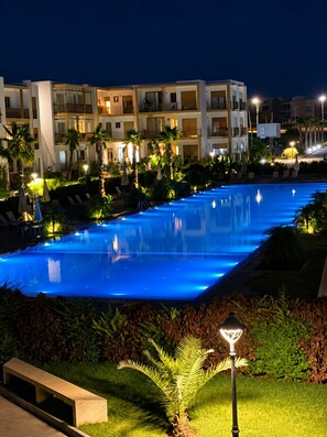 Ein Swimmingpool mit blau beleuchtetem Rand, umgeben von Palmen und Liegestühlen, mit einem Hotelgebäude im Hintergrund.