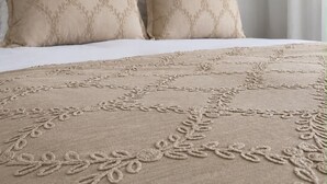 Ein Bett mit einer beigen Bettdecke und passenden Kissen, einem Kopfteil und einem Fenster mit Vorhängen.