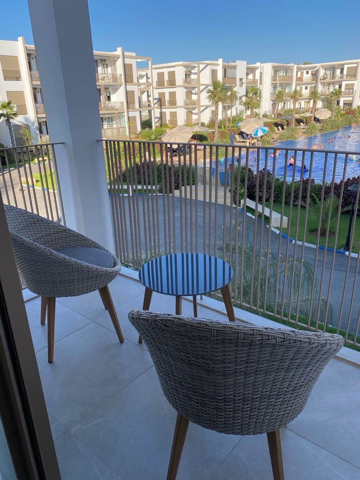 A balcony with wicker chairs, a pool area with umbrellas, and a view of a residential building.