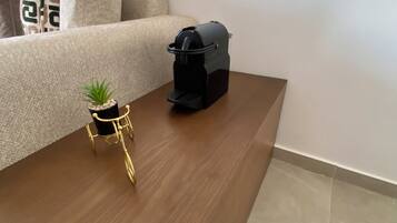Une table basse en bois, sur laquelle se trouve un pot de plante et un objet noir, peut-être un haut-parleur, dans une pièce où se trouvent un canapé beige et des coussins à motifs.