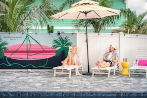 Two people seated on white lounge chairs under a striped umbrella, with a mural of a sailboat and palm trees in the background. - Vilano Beach | Private Pool + Pickleball + Hot Tub (St Augustine)