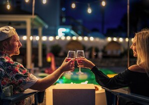 Two people toasting with wine glasses at an outdoor table with string lights in the background. - Vilano Beach | Private Pool + Pickleball + Hot Tub (St Augustine)