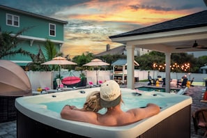 A hot tub with a person reclining, surrounded by outdoor furniture and umbrellas, with a sunset in the background. - Vilano Beach | Private Pool + Pickleball + Hot Tub (St Augustine)