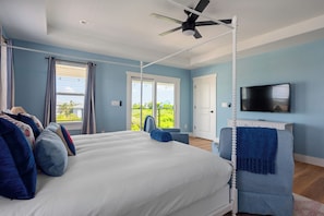 A bedroom with a large bed, a ceiling fan, a TV, and a view of greenery outside. - Vilano Beach | Private Pool + Pickleball + Hot Tub (St Augustine)