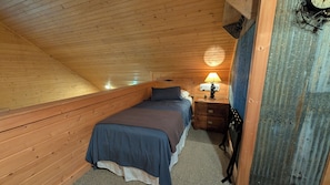 Une chambre avec un plafond en pente, un lit recouvert d’une courtepointe bleue, une table de chevet avec une lampe et un élément de décoration rustique.