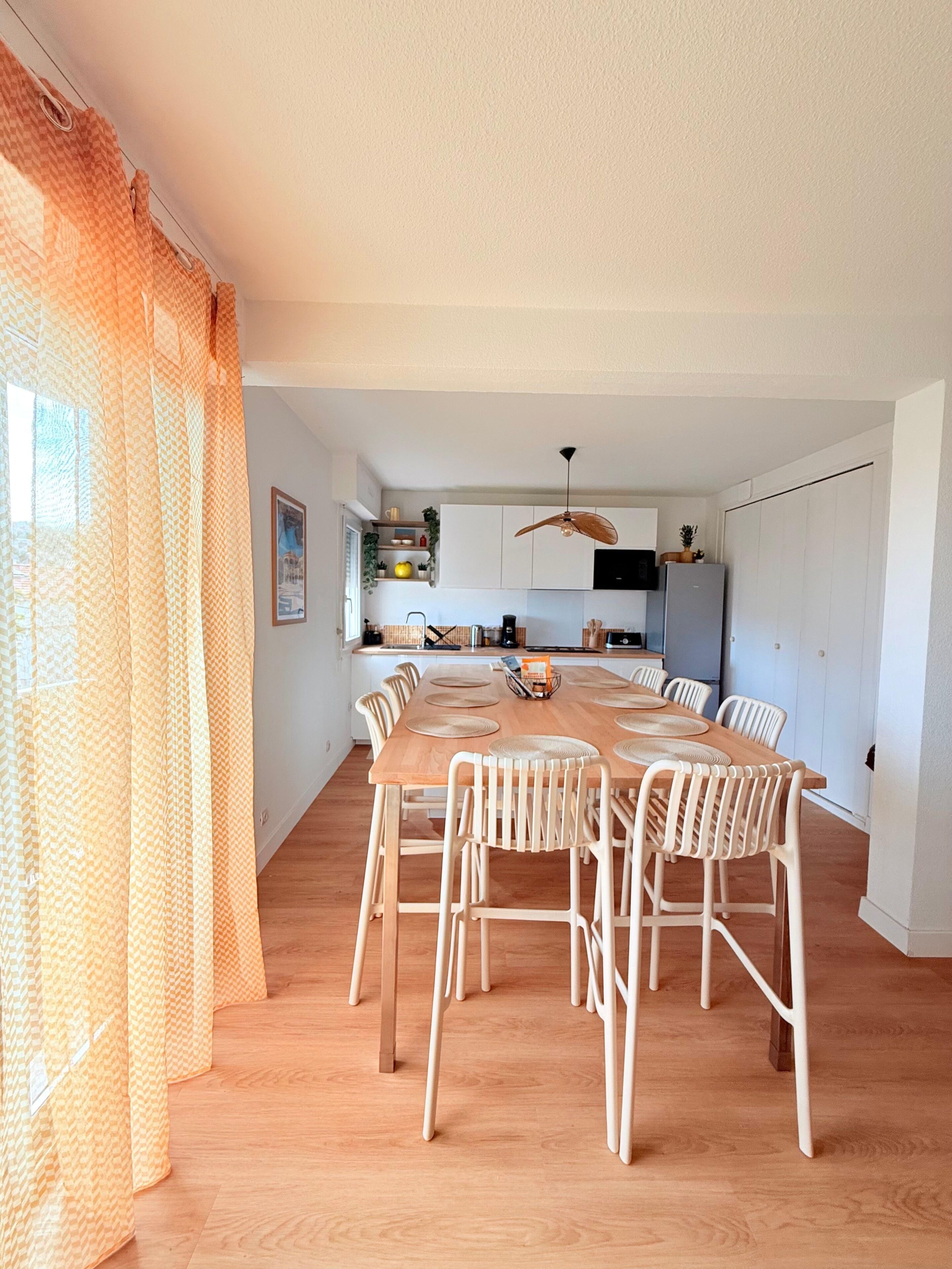A dining area with a wooden table and white chairs, a kitchen in the background, and a ceiling fan.