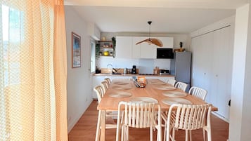 A dining area with a wooden table and white chairs, a kitchen in the background, and a ceiling fan.