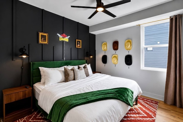 A bedroom with a green upholstered headboard, a bed with white bedding, a red and white patterned rug, a ceiling fan, and a wall with framed pictures and hats.
