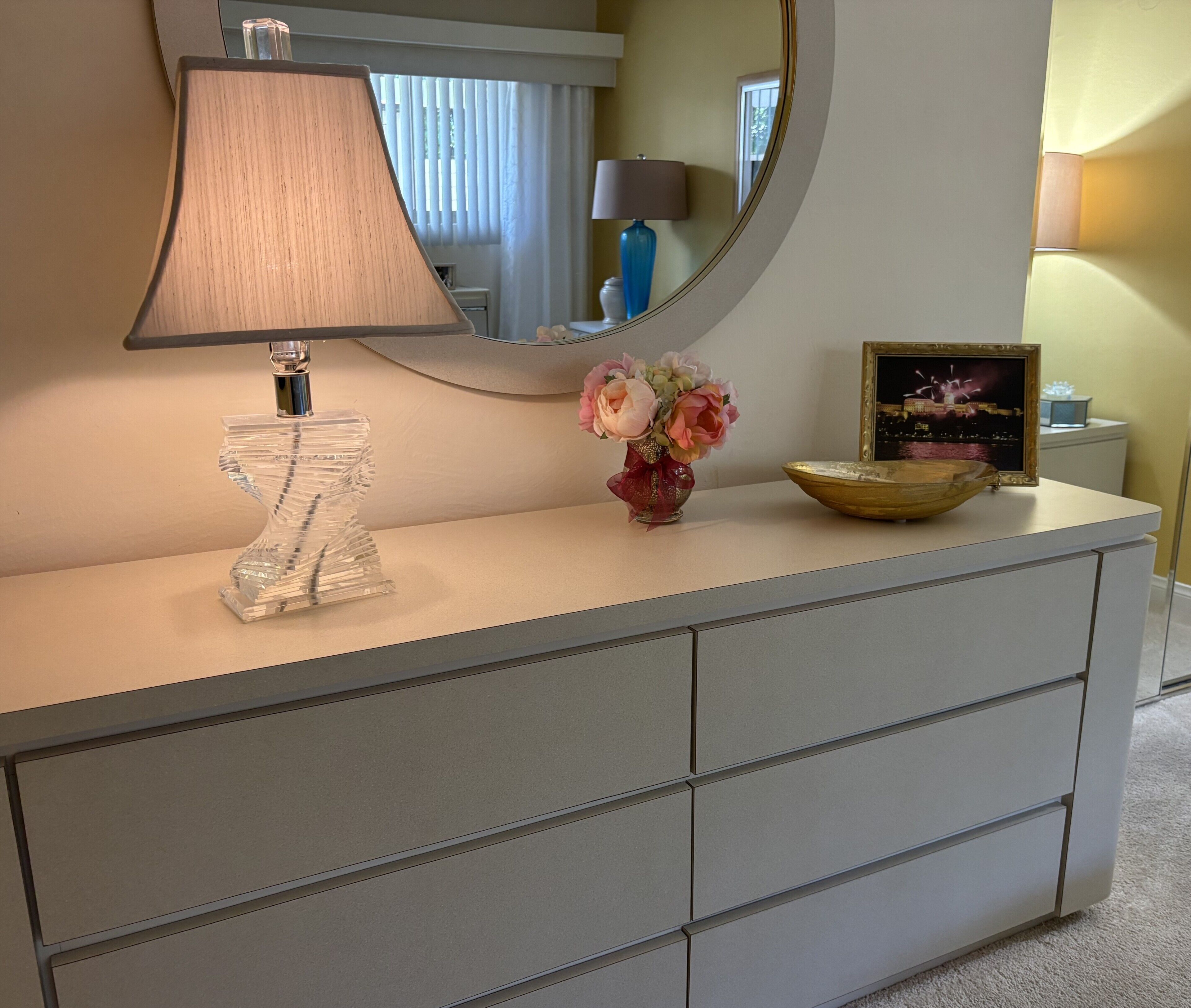 Un meuble-lavabo moderne pour salle de bain, agrémenté d’un miroir, d’une sculpture en verre transparent, d’un vase avec des fleurs et d’un tableau encadré.