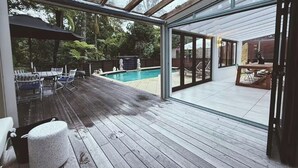 An outdoor deck with a pool, wooden flooring, and glass doors leading to an interior space. - Luxury Coastal Home l Pool, Spacious Entertaining Areas l Steps to the Beach (North Shore)