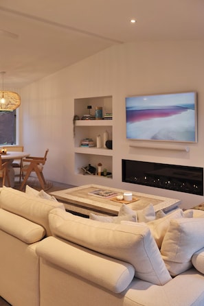 A modern living room with a sofa, a coffee table, a fireplace, and a large TV mounted on the wall. - Luxury Coastal Home l Pool, Spacious Entertaining Areas l Steps to the Beach (North Shore)