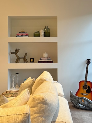 A living room with a sofa, a guitar, and a bookshelf with books and decorative items. - Luxury Coastal Home l Pool, Spacious Entertaining Areas l Steps to the Beach (North Shore)