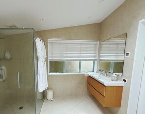 Un bagno moderno con cabina doccia in vetro, un accappatoio bianco appeso, una finestra con tende e un mobile bagno in legno con specchio.