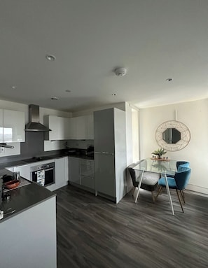 A modern kitchen with white cabinets, a stainless steel range hood, and a dining area with a glass table and blue chairs. - Stylish 2BR | Parking | Near Shops/Station/Parks (Edgware)