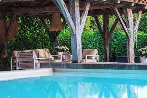 Een zwembadterras met een houten pergola, ligstoelen en een tafel met een bloevas.