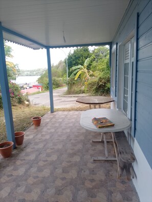 Une terrasse couverte avec une table ronde et des chaises, des plantes en pot et une vue sur une route et la verdure environnante.