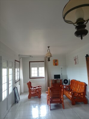 Une pièce aux murs blancs, avec un ventilateur de plafond, un téléviseur et deux chaises en bois.
