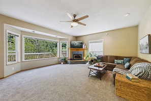 A spacious living room with a fireplace, a ceiling fan, and large windows offering a view of greenery. - Spacious 4-bedroom house in peaceful Eureka with backyard and fire pit (Eureka)