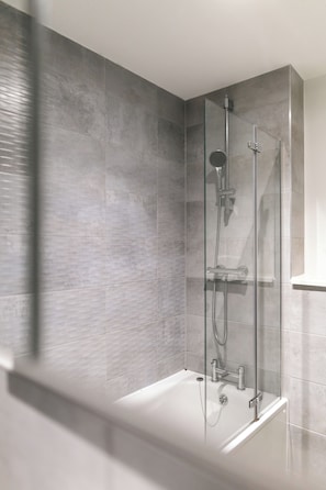 Een moderne badkamer met een douchecabine van glas, een witte badkuip en grijze betegelde wanden.