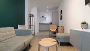 En moderne stue med en sofa, et rundt bord og en stol. Der er et lille tekøkken i baggrunden med en vask og skabe.