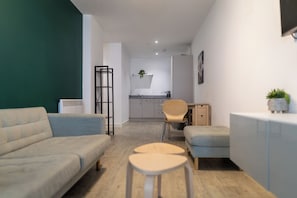 Een moderne woonkamer met een bank, een klein rond tafeltje en een stoel. Achterin is een kitchenette met een spoelbak en kastjes.