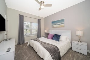 Une chambre à coucher avec un lit, une table de chevet, une télévision, un ventilateur de plafond, une fenêtre avec des rideaux et un tableau au mur.